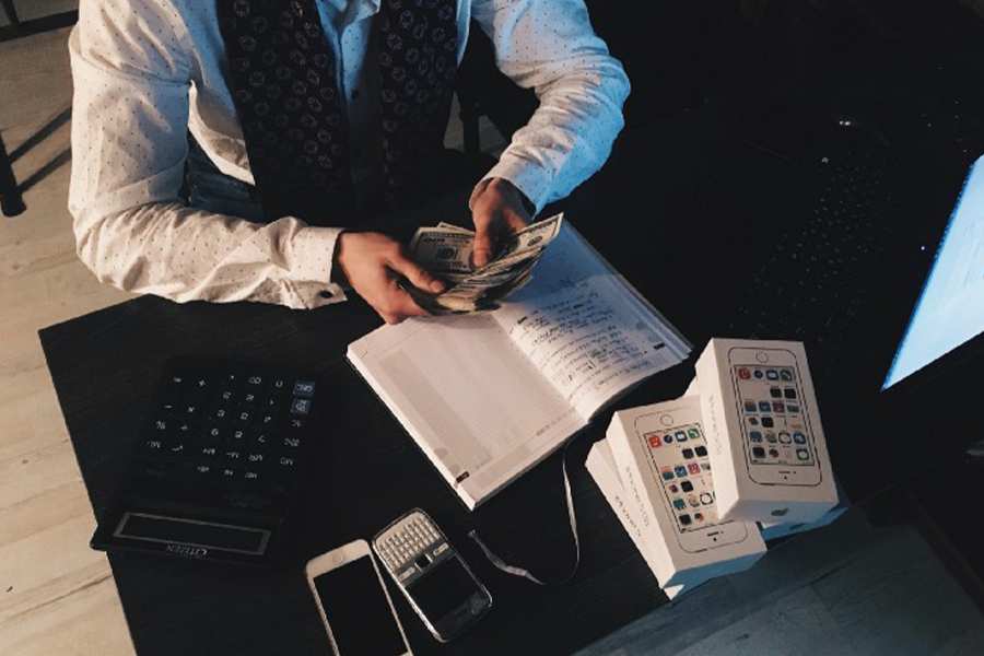 Man holding a cash while counting it with a caluclator, notebook, mobile phone and a box in front of him.