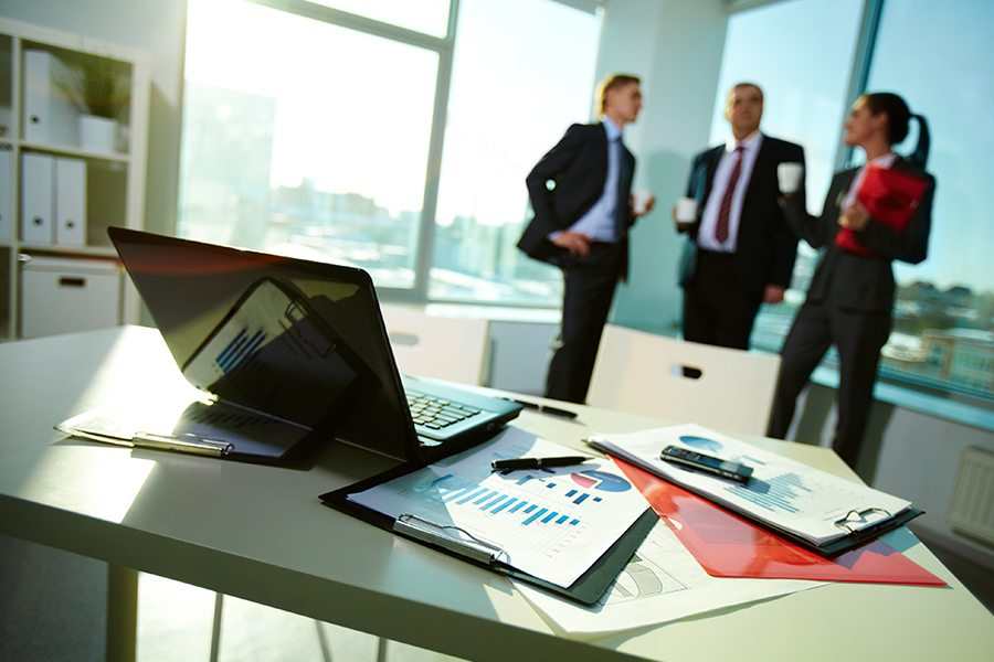 Image of business documents on workplace with three partners interacting on background.