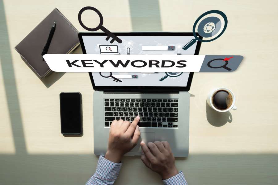 Man's hand searches for keywords on his laptop with coffee, phone, and magnifying glass in the background.