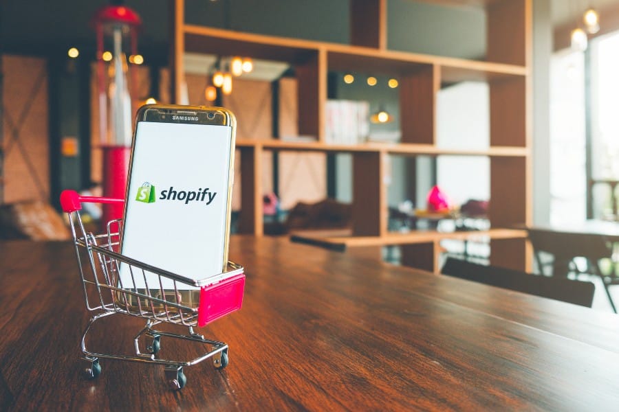 Shopify app on Mobile Phone screen and in a shopping cart on wooden table.
