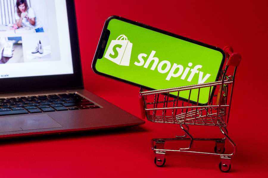 Mobile phone with Shopify logo in a small grocery cart along with a laptop on the left side.