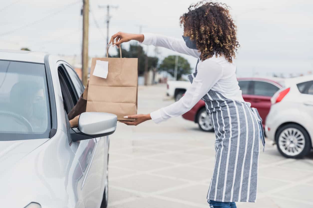 Restaurant worker with protective face mask giving order bags to customer in car.