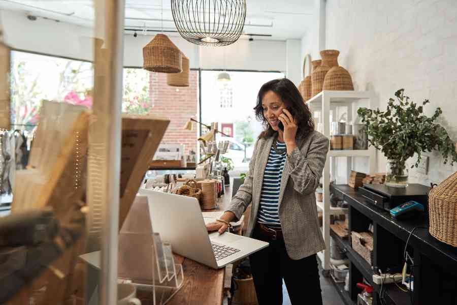 Smiling young Asian woman standing behind a counter in her stylish boutique working on a laptop and talking on a cellphone.