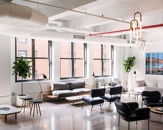A coworking space with big windows, exposed pipes, and black, white, and gray seating furniture.