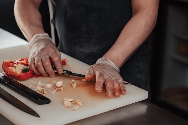 A cook wearing gloves chops garlic and bell peppers on a white cutting board.