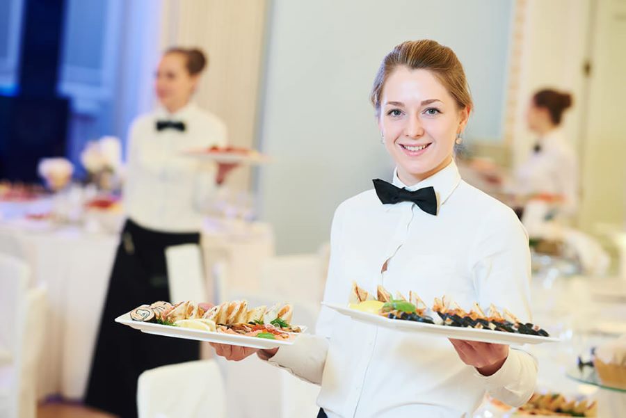 Three female servers in white shirts with black bow ties carrying trays of food in a banquet hall.
