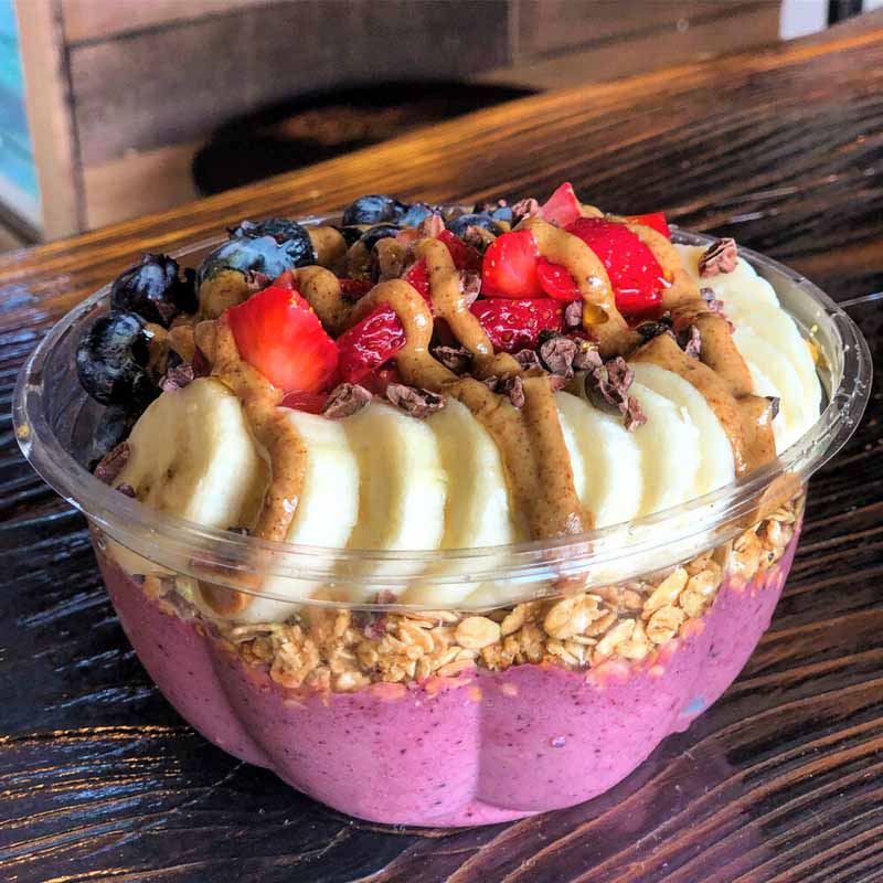 An acai bowl in a plastic container topped with granola, bananas, blueberries, strawberries, and peanut butter.