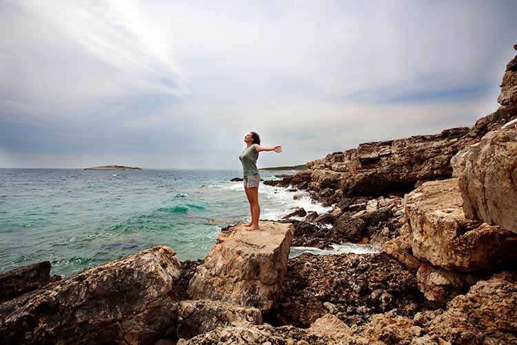 A barefoot woman in shorts and a t-shirt standing on a rocky shoreline, with waves hitting the rocks, raising her hands, and breathing in the salty ocean air.