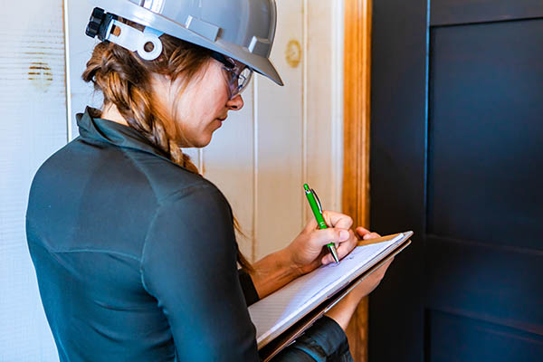 A woman home inspector in a hardhat with a clipboard taking notes during her inspection.