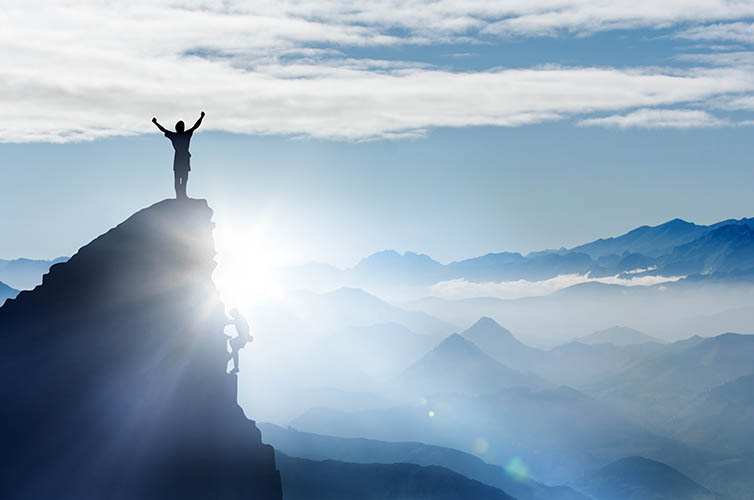 A woman standing on the pinnacle of a mountain overlooking mountains in the fog below, raising her hand up to the sky as she contemplates her future.