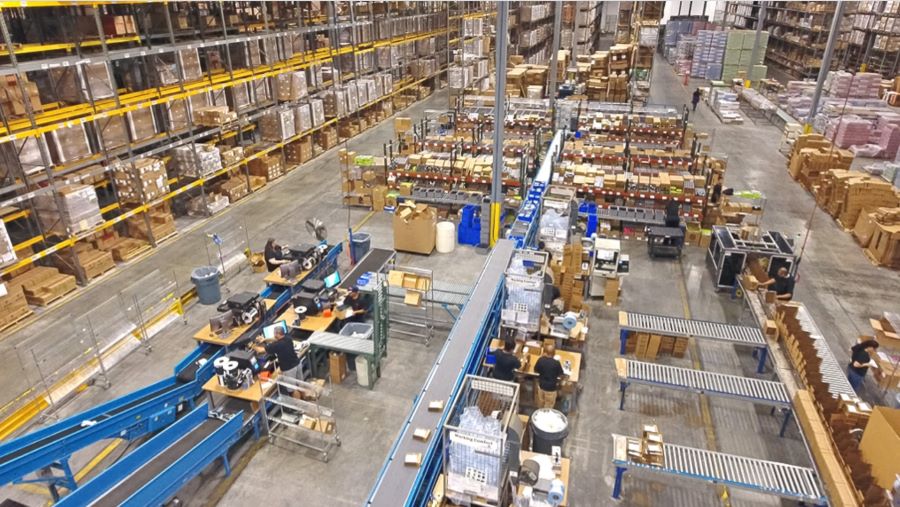A large fulfillment center with boxes stored on floor-to-ceiling shelving next to additional boxes on conveyor belts and packing stations.