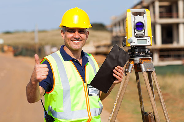 land surveyor wearing a yellow hard hat and safety vest standing next to surveying equipment and giving the thumbs up.