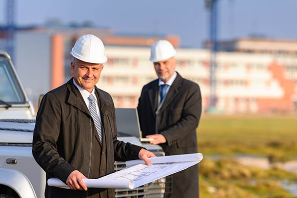 Two men in hardhats reading blueprints outdoors.