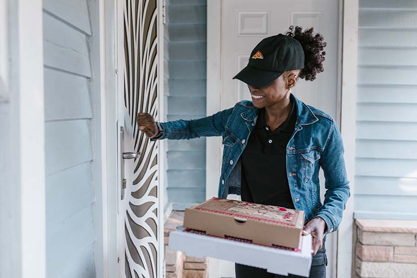 Female delivery driver holding pizza boxes knocking on door of a house.