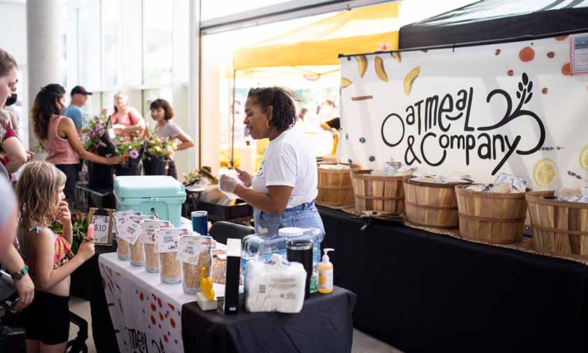 A person standing between a table of oatmeal packages and a sign reading Oatmeal & Company interacting with customers.