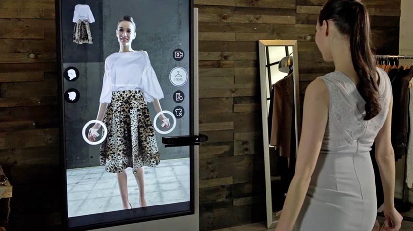 An in-store shopper tries on an outfit virtually by standing in front of a smart mirror with an embedded camera.