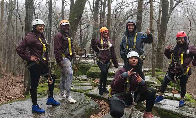 Six people in harnesses and outdoor gear giving thumbs-up to the camera as they prepare for a zipline adventure.