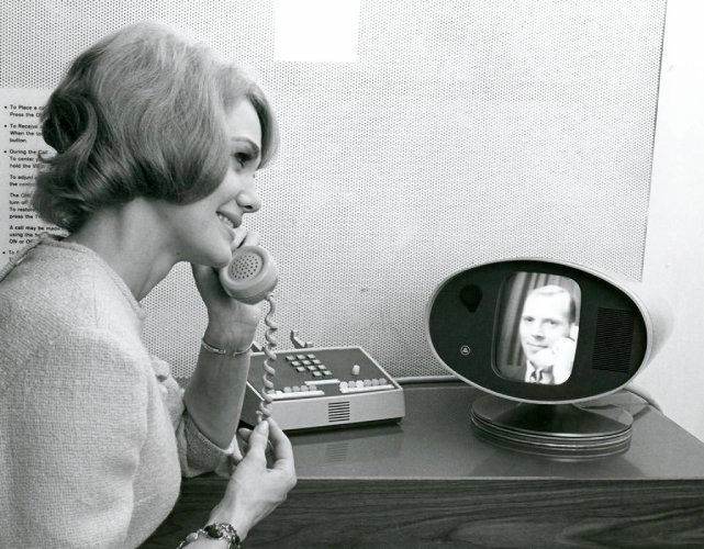 A woman who’s holding a telephone to her ear looks at the Picturephone screen, seeing the person at the other end of the line.
