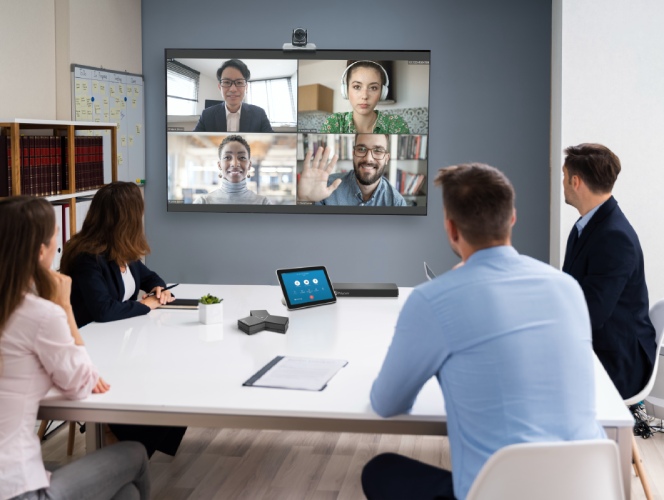 A hybrid team meeting with four people in a physical room looking at a television screen that displays four video meeting participants.