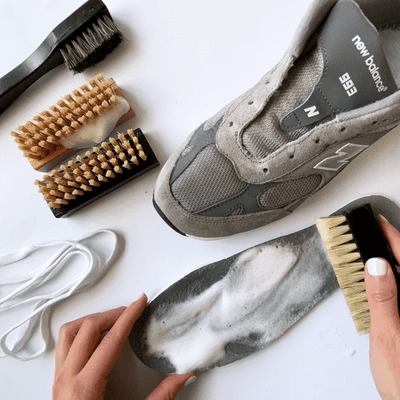 A person cleaning a gray sneaker insole with a soapy brush, surrounded by shoe-cleaning brushes and white laces.