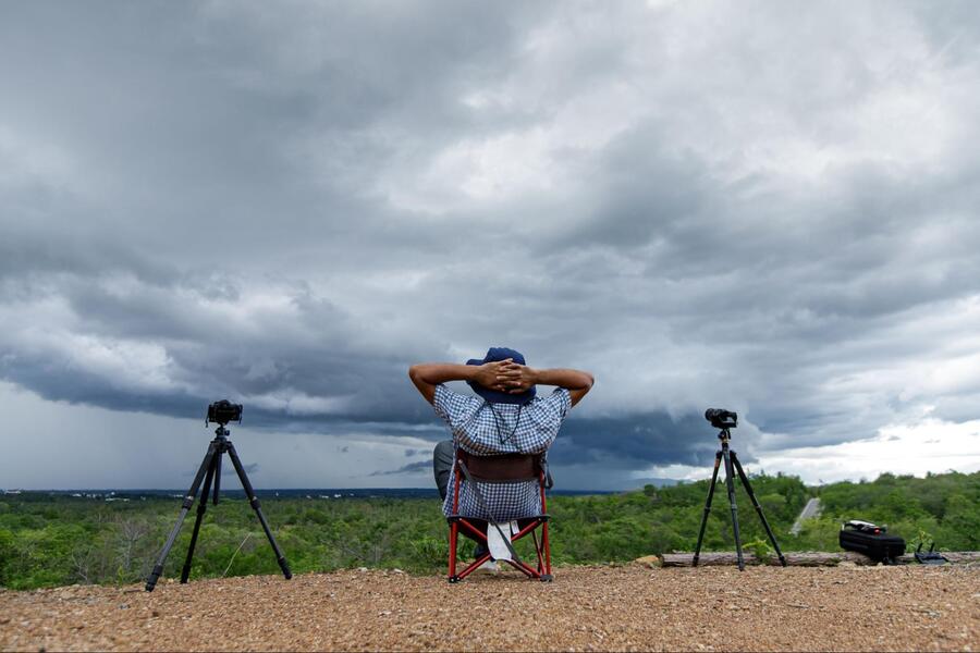 A man sits in a folding chair on a hilltop, hands behind his head, watching storm clouds roll in, with two cameras on tripods.