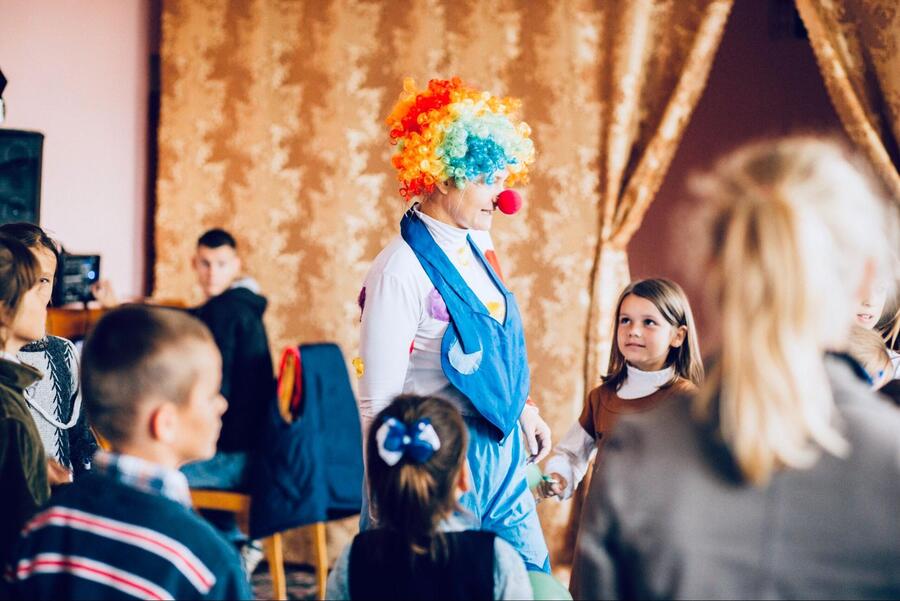 A clown in a rainbow wig and blue overalls entertains a group of children indoors, engaging them in conversation and play.