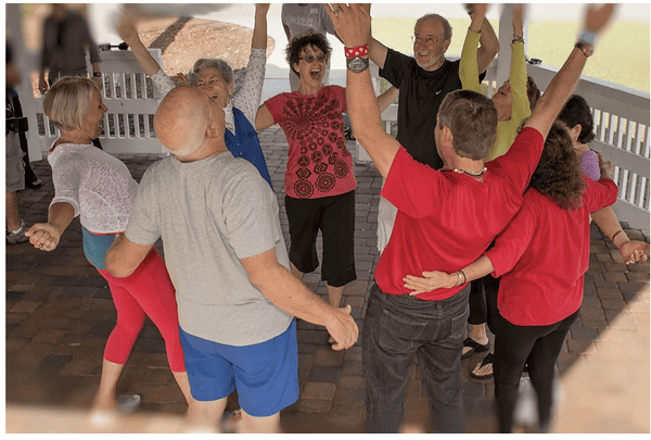 A group of older adults gathers in a circle under a pavilion, laughing and raising their arms in a lively yoga session.