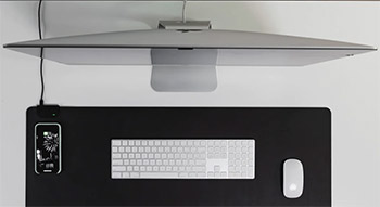 A large Apple monitor and a wireless desktop charging pad with a charging iPhone, wireless keyboard, and mouse on top.