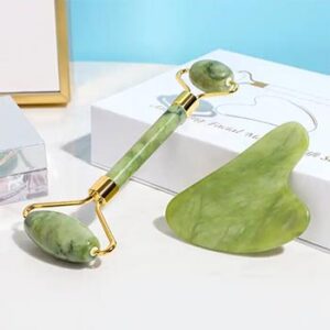 A jade facial roller with a wheel on either end and a jade gua sha tool leaning against a white box.