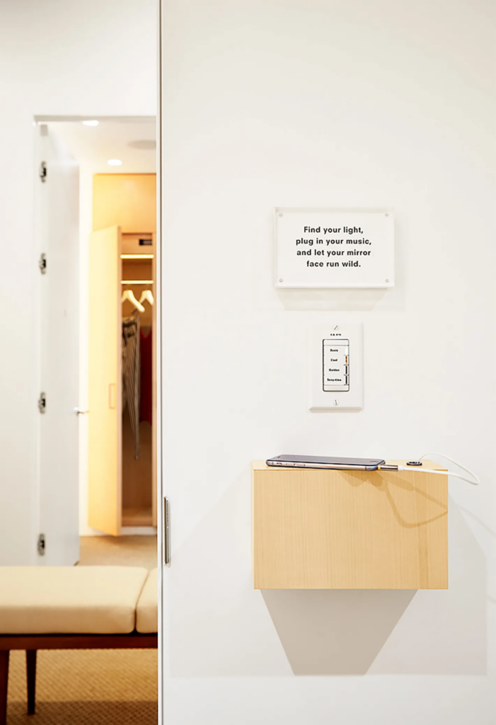 Fitting room scene with a wall-mounted wooden shelf holding a smartphone connected to a charging cable. Above the shelf is a light control panel with options labeled 'Warm,' 'Cool,' 'Outta,' and 'Sexytime.' A sign above reads, 'Find your light, plug in your music, and let your mirror face run wild.' An open closet with clothes and a bench are visible in the background. Reformation store fitting room design