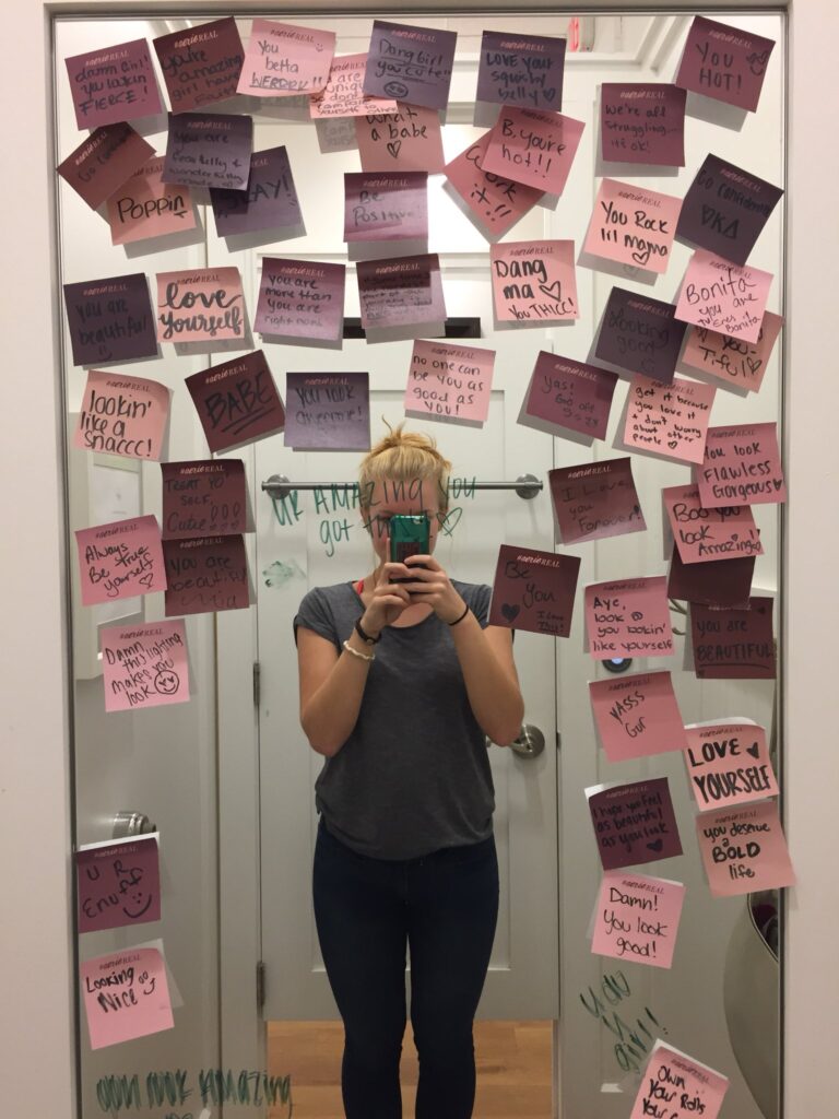 Mirror covered with colorful sticky notes displaying handwritten body-positive and self-love messages like 'You are beautiful,' 'Love yourself,' and 'You rock!' A woman stands in front of the mirror taking a photo, partially visible behind the notes.Aerie fitting room design