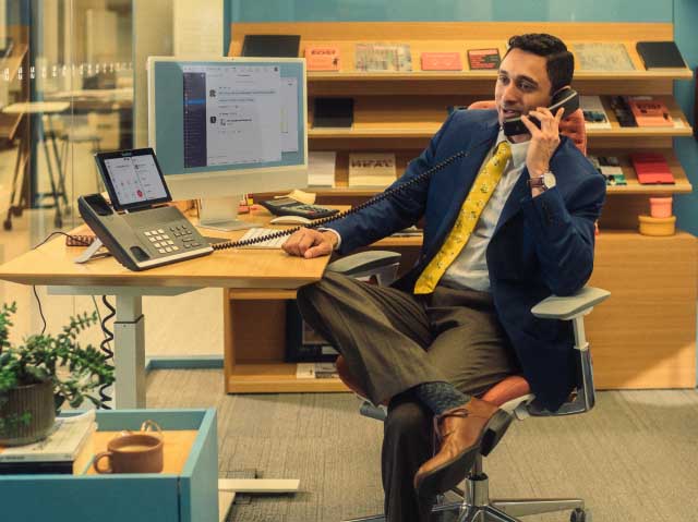 A man sitting on a chair and talking on the phone placed at the top of a computer desk.