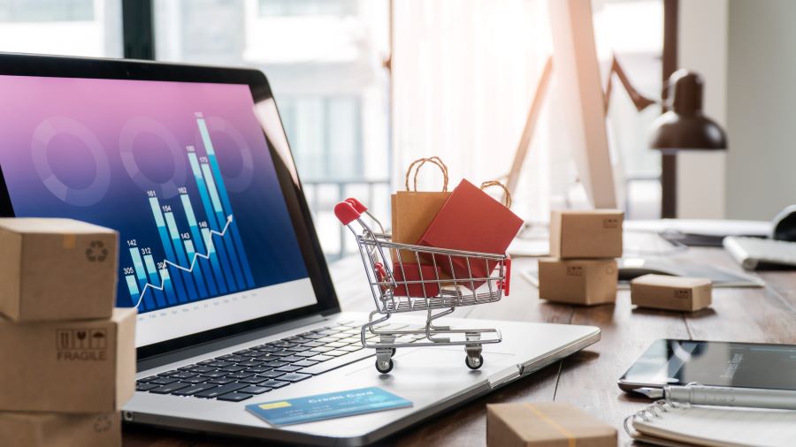 Image showing shopping bags in a cart, a credit card on a laptop, paper boxes on a table, and a sales data economic growth graph on the screen. Depicts buying and selling services online, e-commerce concept.