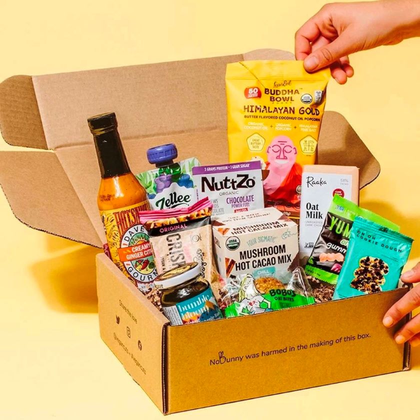 Open subscription box full of snacks on a yellow background with a hand reaching in to grab popcorn. 