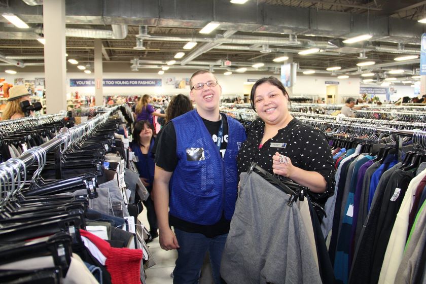 Two Goodwill employees smiling and standing in an aisle of clothing while holding an item of clothing on a hanger.