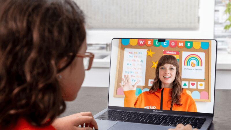 A young person looking at a computer screen showing a video call with a teacher waving in front of a colorfully decorated cork board.