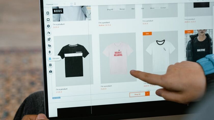 A hand pointing at a computer screen showing an online store with product listings for un-printed tee shirts.