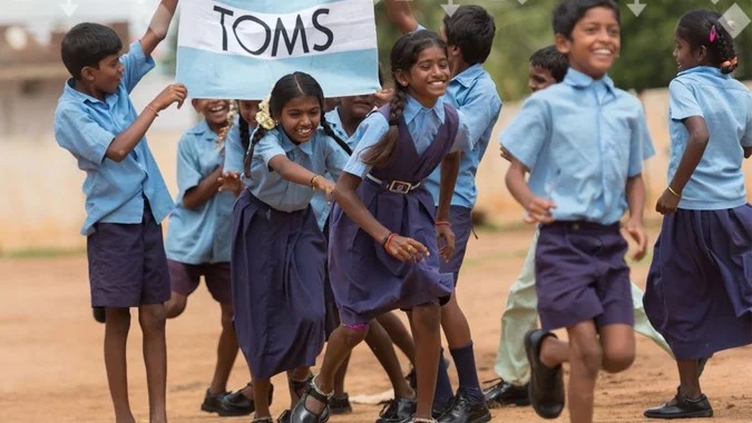 a group of children in school uniforms smiling and running with a sign that reads "TOMS".