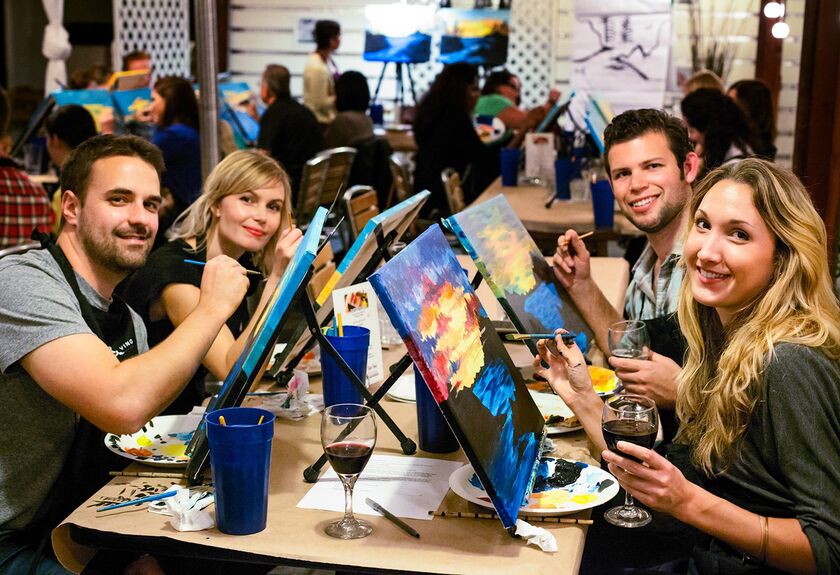 a group of smiling people sitting at a bench with a painted canvas and a glass of wine in front of each person.
