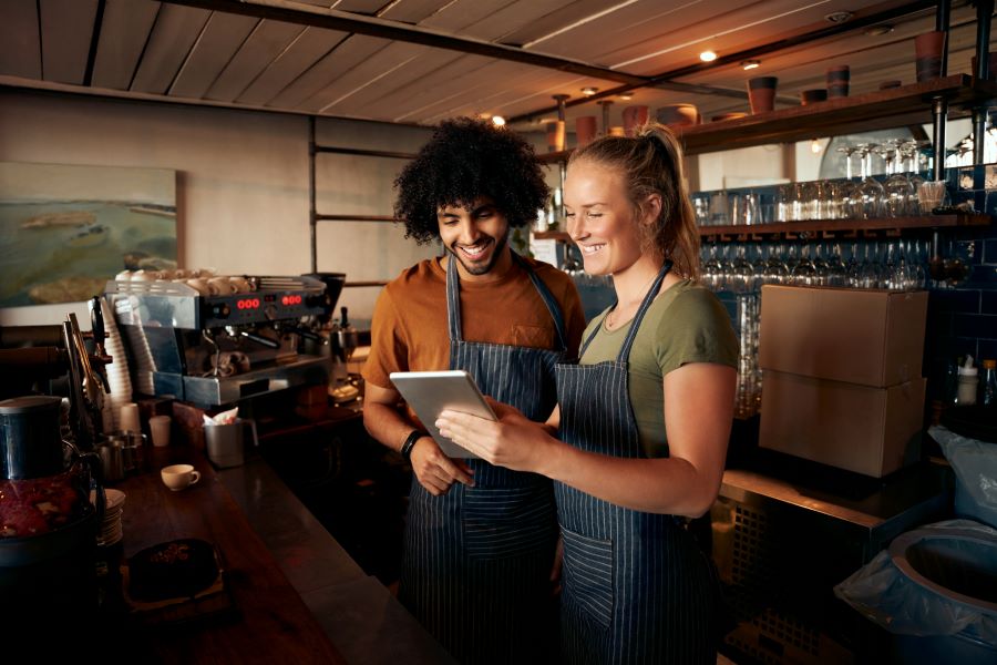Smiling waiter and waitress wearing apron using digital tablet standing behind counter in cafe.
