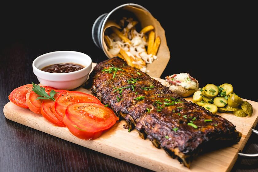 Rack of ribs on a cutting board surrounded by sliced tomatoes, pickles, french fries and a ramekin of sauce.