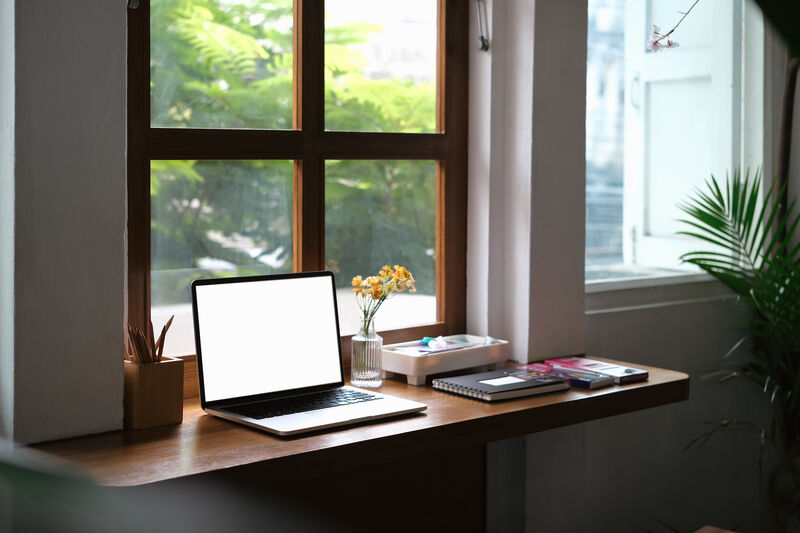 Laptop with blank display, books and stationery on wooden desk near window.