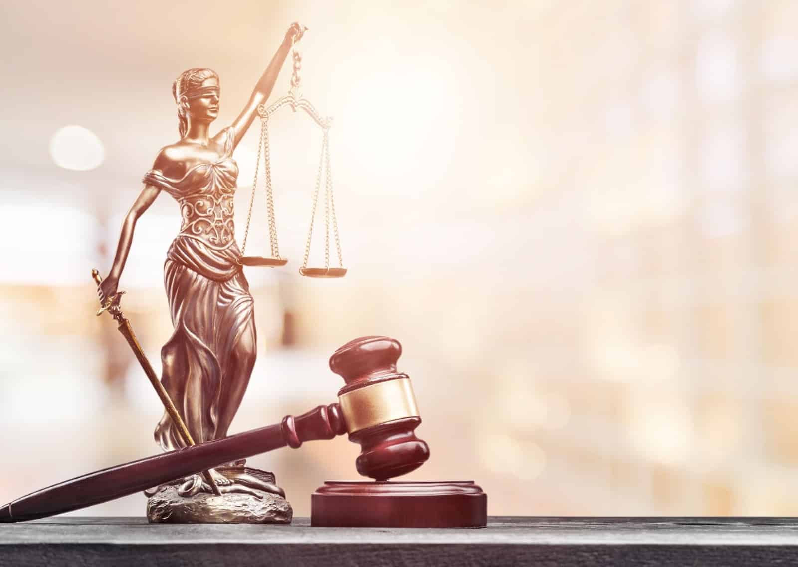 Image of small brass statuette of the Goddess of Justice holding scales and sword, and gavel sitting in foreground over blurred, bright background.
