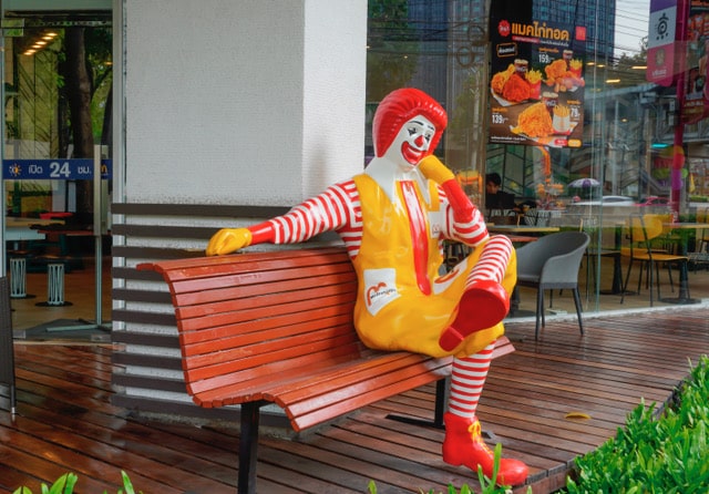Statue of Ronald McDonald, sitting on bench in front of McDonald's restaurant.