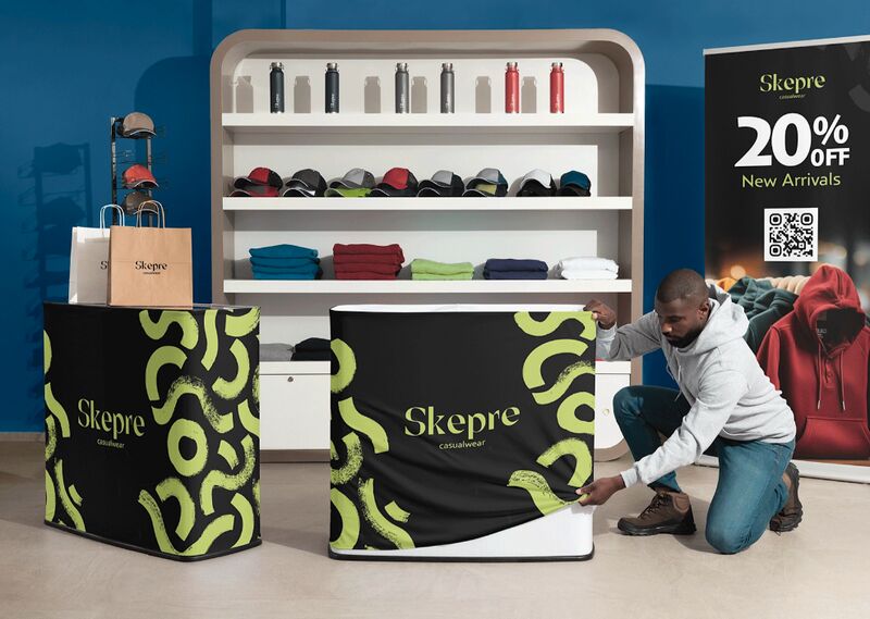 A portable VistaPrint podium with stretch fabric branding being placed on a fold-away podium by an African-American man in a gray sweatshirt in front of a trade booth with clothing on display shelving behind him.