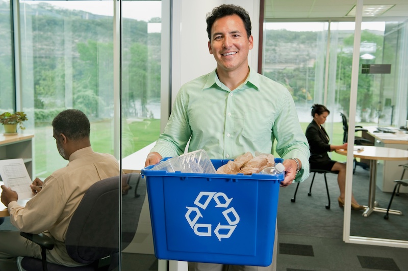 A smiling male office employee in light green shirt in a glass walled office, taking out a full blue recycling bin.