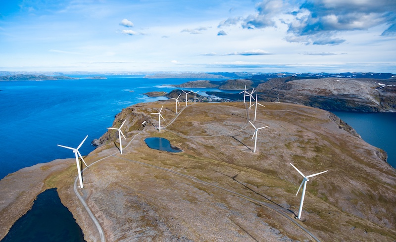 Aerial of a windfarm with 12 turbines on hilltop overlooking a large fjørd.