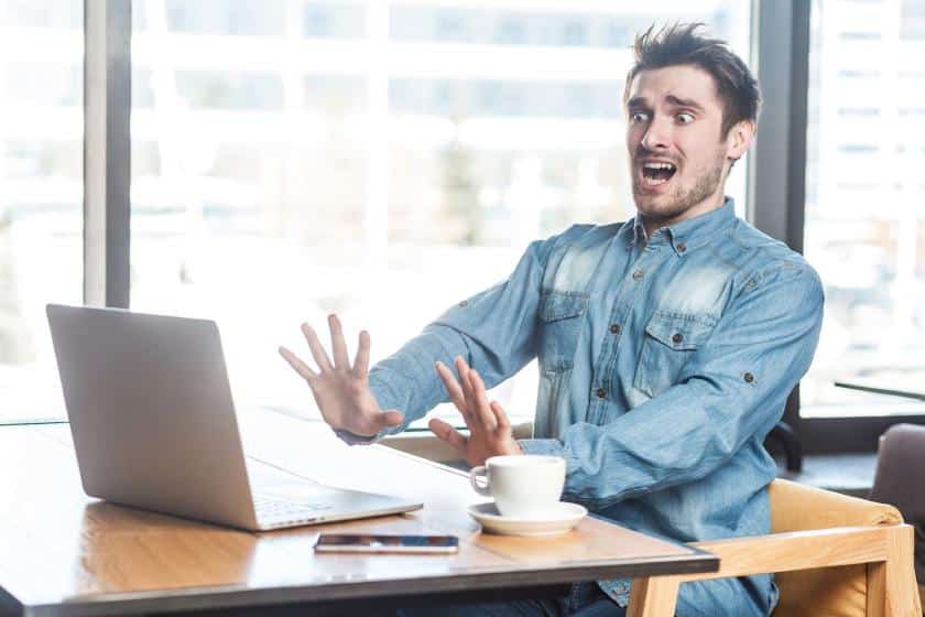 Young man sitting in cafe with a scared face while screaming at a laptop because of a mistake.