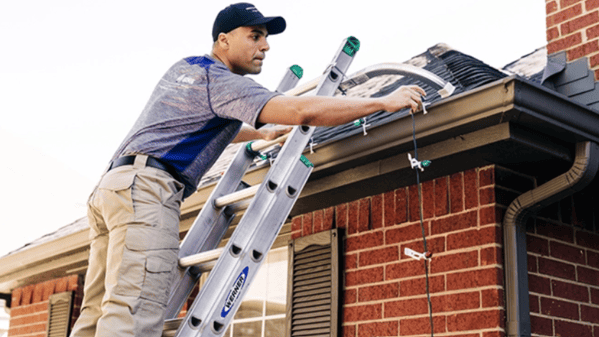 Man on ladder hanging Christmas lights along a roofline