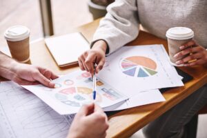 Close up of two colleagues discussing their work over wooden desk. They are studying charts. Woman holds a pen and a cup of coffee. Casual style. Concept of success